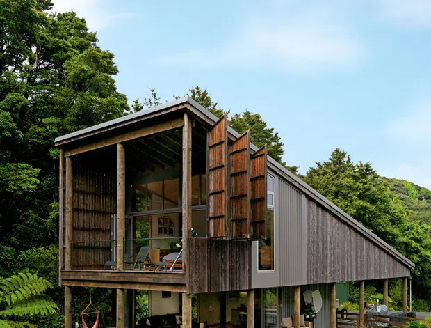 A contemporary elevated house set within a lush, wooded landscape. The building features a long sloping roof, extensive weathered timber screening and exposed timber posts, with an open-sided lower level and a glazed upper living area. Native-style planting, ferns and lawn surround the structure.