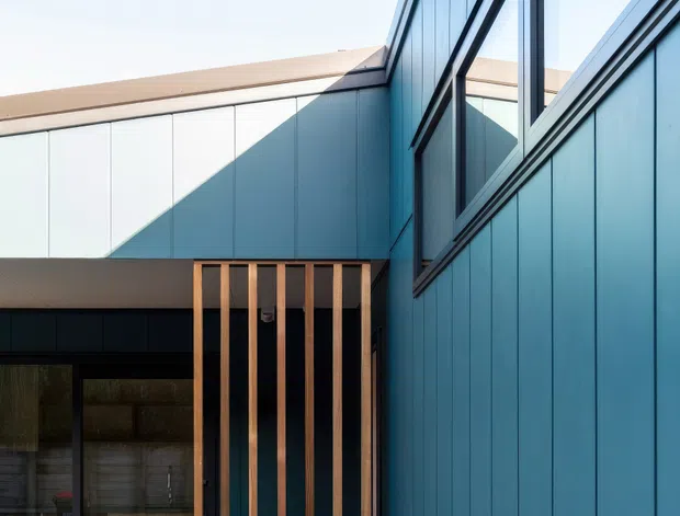 Close-up exterior architectural view of a contemporary building corner with blue vertical-panel cladding, dark-framed floor-to-ceiling glazing, and a timber-slatted screen over an entry or deck. Clean geometric forms and strong sunlight create angular shadows across the facade.