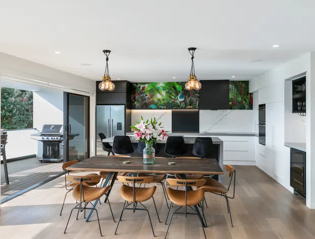 Open-plan dining and kitchen interior with a large dark timber dining table surrounded by sculptural wooden chairs, facing a contemporary kitchen with white cabinetry, dark overhead joinery, marble-look splashback, and vibrant botanical artwork. Pendant lights hang above the table, while full-height glazing and an open door connect the dining area to an outdoor deck with a barbecue.