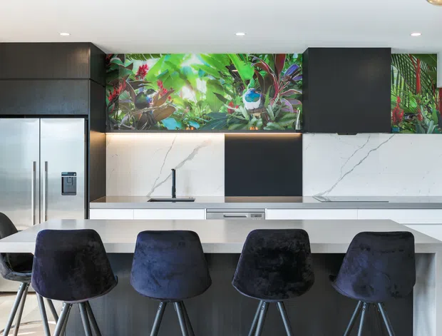 A contemporary kitchen with a large white island and four dark upholstered bar stools. The space features white veined stone surfaces, matte dark cabinetry, integrated stainless-steel appliances, under-cabinet lighting, and a vivid tropical botanical splashback artwork or panel.