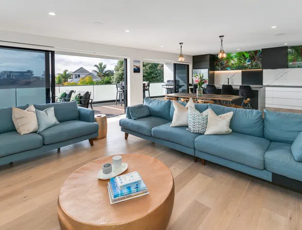 Bright open-plan living area with a large blue sectional sofa and matching armchair arranged around a round timber coffee table. The space has light timber flooring, white walls and ceiling, expansive sliding glass doors opening to a balcony or outdoor area, and a contemporary kitchen with dark cabinetry, white joinery, pendant lights, and a colourful botanical artwork backsplash.