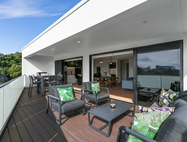 Contemporary rooftop or elevated terrace with dark timber decking, glass balustrades, outdoor dining and lounge furniture, and wide sliding glass doors opening into a bright open-plan interior. The white flat-roofed facade and generous glazing create a clean, modern indoor-outdoor connection.