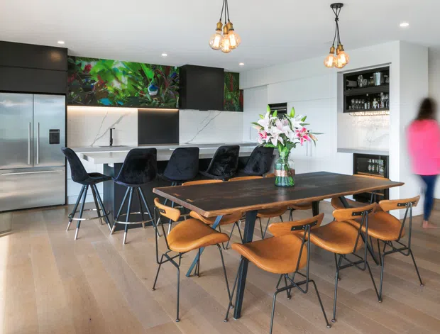 Bright open-plan kitchen and dining area with a large dark timber dining table, sculptural wooden chairs, a contemporary black kitchen island with stools, stainless-steel double-door refrigerator, dark cabinetry, white marble-look splashback, and a built-in display and bar area. Decorative pendant lights and a colourful botanical mural add visual interest.