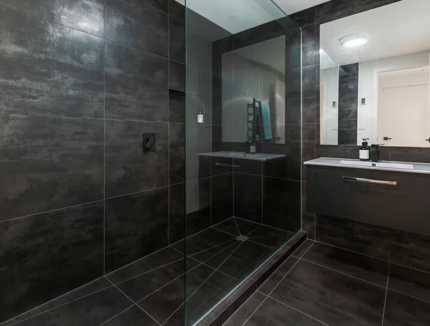A contemporary bathroom with a large frameless glass walk-in shower, dark charcoal stone-look wall and floor tiles, and a floating dark vanity with a white benchtop and integrated basin. A large mirror, recessed shower fittings, and minimalist black fixtures create a sleek, moody interior.