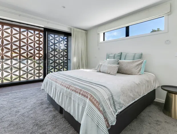 Bright contemporary bedroom with a low-profile upholstered bed dressed in layered white and pale blue-grey bedding. The room has soft grey carpet, white walls and ceiling, sheer curtains, and a long horizontal clerestory window. Full-height glazed doors feature a prominent geometric patterned privacy screen, while a small metallic side table sits beside the bed.