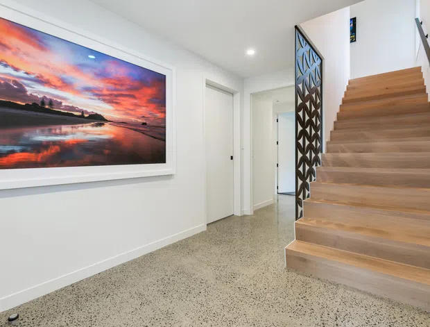 Bright contemporary residential hallway and stairway with white walls, a polished speckled terrazzo floor, and a broad light timber staircase. A decorative dark geometric screen forms the stair balustrade, while a large framed landscape artwork provides a colourful focal point.