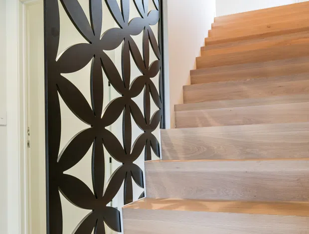Bright contemporary stairway with light timber floating-style treads and risers, paired with a dark decorative metal screen featuring a repeating organic floral cut-out pattern. White walls and a terrazzo-like floor are visible beside the stair, with a doorway beneath or adjacent to the stairs.