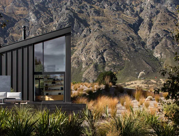 Exterior view of a compact contemporary dark-clad building set against a dramatic rocky mountain landscape. The facade features vertical dark panels, extensive floor-to-ceiling glazing, a sharply sloping roofline, and a small outdoor seating area. Native grasses and sculptural landscaping surround the building, while the interior shelving and furnishings are visible through the glass.