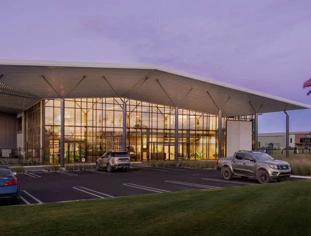A contemporary commercial building viewed from across a landscaped lawn and parking area at dusk. The main facade features a broad, shallow projecting roof canopy, extensive floor-to-ceiling glazed curtain walling, and dark metal cladding. Interior lighting glows through the transparent entrance or lobby volume. Flagpoles and parked vehicles are visible outside, but the building is the dominant subject.