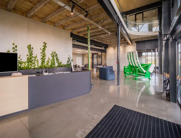A spacious contemporary commercial reception and lobby area with an open-plan industrial character. The space features a polished concrete floor, exposed timber ceiling structure, dark steel beams and framing, full-height glazing, a large reception desk, indoor planting, and a bright green sculptural lounge chair. An upper-level mezzanine or balcony is visible at the rear.