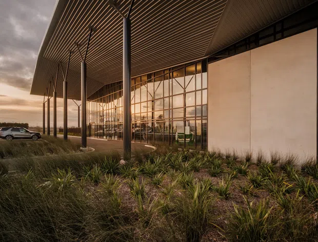 Exterior architectural photograph of a contemporary commercial building with a dramatic deep projecting roof canopy, slim dark structural columns, extensive floor-to-ceiling gridded glazing, and a large pale rendered wall. Native-style landscaped planting surrounds the building, with a vehicle visible in the distance. The warm sunset light and reflective glazing create a refined, atmospheric presentation.