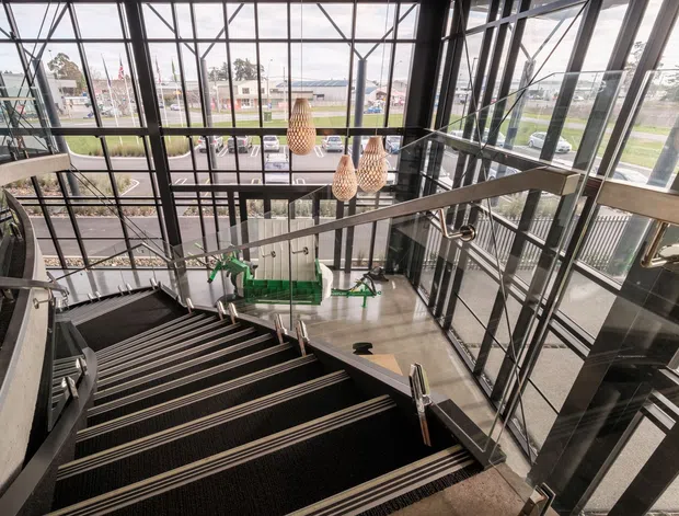 A dramatic multi-level interior stairway viewed from above, featuring broad dark carpeted stairs with metallic edge trims, stainless-steel handrails, extensive black-framed floor-to-ceiling glazing, exposed structural framing, and several woven pendant lights. A lower-level lobby with green seating or display elements is visible below, while streetscape and landscaping can be seen through the glass.