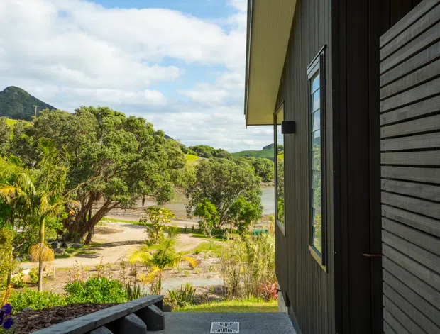 Exterior view of a contemporary dark-clad building, photographed from a covered entry or deck. The facade features horizontal timber-like wall cladding, vertical dark cladding, a deep roof overhang, and a tall narrow window. A landscaped garden and rolling green hills are visible beyond the building.