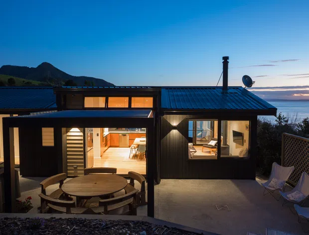 Exterior twilight view of a compact dark-clad coastal home, showing a blue metal roof, illuminated interior spaces through large windows and open doors, and a covered outdoor dining terrace with a round table and chairs. The property overlooks the sea, with mountainous terrain visible in the background.