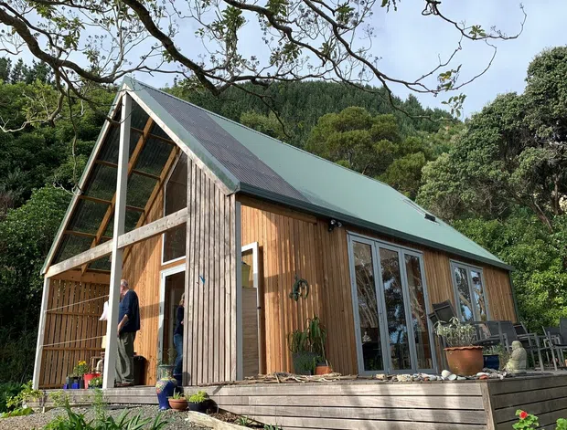 Exterior view of a compact timber-clad cabin or small house set within dense native vegetation. The building has a steep asymmetrical pitched roof with green metal roofing, vertical timber wall cladding, a glazed gable end, large glazed doors and windows, and a raised timber deck with outdoor pots and seating.