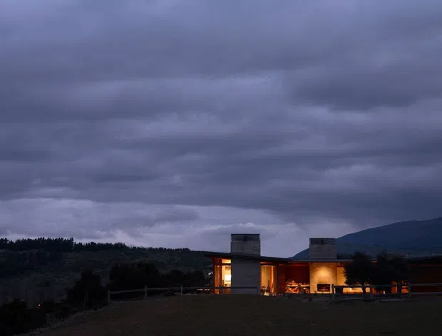 A low, single-storey modern residence photographed at dusk, set against a dark wooded hillside and mountain backdrop. The house has a long horizontal form, broad roofline, warm interior lighting, and two prominent rectangular masonry chimney or tower elements. The scene is dominated by dramatic storm clouds and a moody blue-purple evening atmosphere.