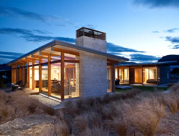 Twilight exterior view of a low-slung contemporary house set within a landscaped, grass-covered site. The home features extensive floor-to-ceiling glazing, warm timber-framed windows and doors, a broad shallow roof, a prominent raw concrete chimney or central wall element with a louvred cap, and interconnected single-storey wings. Warm interior lighting contrasts with the cool blue evening sky.