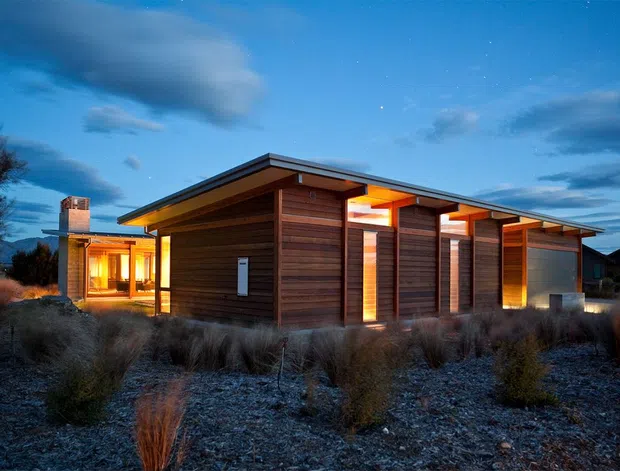 A single-storey contemporary timber-clad house photographed at dusk, with a low-pitched projecting roof, deep eaves, warm illuminated interior spaces, and a rugged landscaped foreground of grasses and gravel. The building has strong horizontal wood siding, timber posts and beams, large glazed openings, and a compact pavilion-like composition.