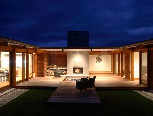 A dramatic twilight view of a low-profile timber-clad courtyard house arranged around a central landscaped courtyard. Warm interior lighting spills through extensive glazed doors, illuminating a raised timber deck, central outdoor dining setting, built-in fireplace, stacked firewood, and a dark vertical chimney or screened feature. The composition is symmetrical and strongly connected to the courtyard.