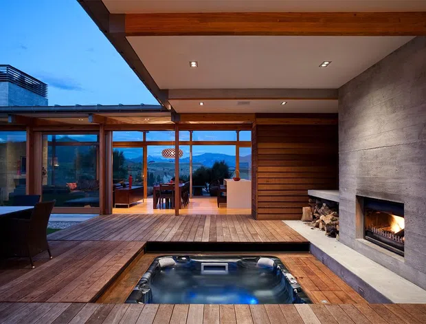 A sophisticated covered outdoor entertaining area with a timber deck, built-in spa, large concrete fireplace, and extensive timber-framed glazing. The space opens toward a view of mountains and landscaped grounds, with an adjoining dining and lounge area visible through the glass. Warm recessed lighting and timber finishes create an inviting evening atmosphere.