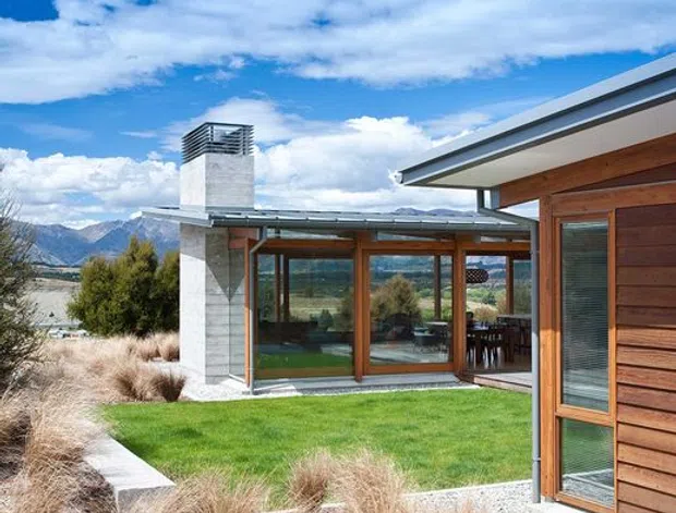 Exterior view of a contemporary single-storey residence set within a landscaped, rocky site. The building combines horizontal timber-clad walls, extensive timber-framed glazing, broad roof overhangs, and a prominent pale concrete chimney or vertical feature. A lawn courtyard is bordered by gravel, ornamental grasses, large rocks, and low concrete edging, with mountain scenery visible beyond.