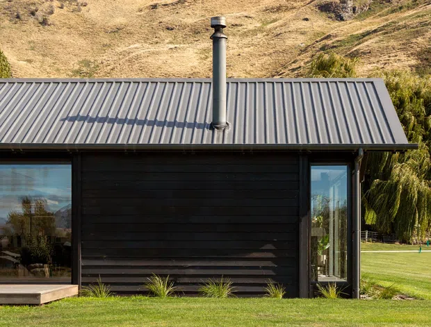 Exterior view of a compact contemporary single-storey building set within a landscaped grassy site and surrounded by steep dry hills. The structure has dark horizontal timber cladding, extensive glazing, a simple grey corrugated metal pitched roof, and a prominent metal chimney, with a small raised deck along the glazed frontage.