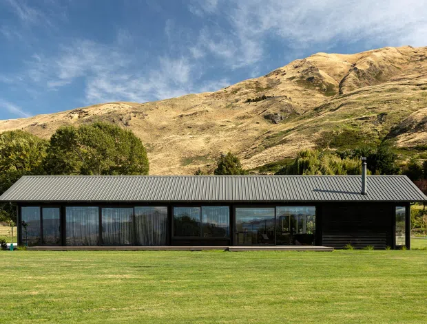 A long, single-storey contemporary house set in a broad lawn beneath a rugged alpine mountain landscape. The building has a simple low-profile rectangular form, dark horizontal cladding, extensive floor-to-ceiling glazing, and a grey corrugated pitched roof with a slender chimney. Mature trees and landscaped grounds surround the home.
