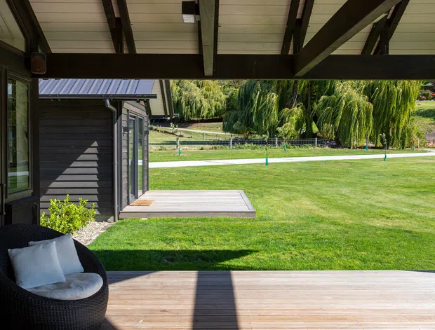 Covered outdoor deck or veranda opening onto a broad manicured lawn, with dark timber posts and beams, a pale lined ceiling, timber decking, and a black woven lounge chair. Glazed doors and part of the house are visible at left, while a small dark-clad outbuilding and landscaped grounds extend beyond the deck.