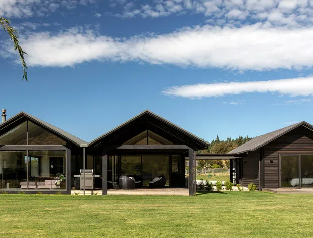 A low-rise modern rural residence composed of three connected dark timber-clad pavilion forms arranged across a broad manicured lawn. The central and left wings have prominent pitched gable roofs with deep overhangs and extensive floor-to-ceiling glazing, while the right wing is a smaller enclosed timber-clad volume with a pitched roof and large window. The setting includes open grass, distant planted trees and a clear sky. The architecture has a restrained contemporary cabin or pavilion character with strong indoor-outdoor transparency.