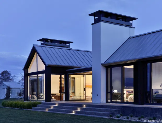 Exterior dusk view of a contemporary residential house with steep standing-seam metal gable roofs, broad floor-to-ceiling glazing, dark window and door frames, and a prominent white rendered chimney with a dark cap. Illuminated interiors are visible through the glazing, including living and dining areas. The home sits on a landscaped lawn with broad entry steps and recessed stair lighting.