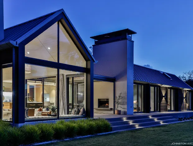 Twilight exterior view of a contemporary single-storey residence with a steep gabled glazed pavilion, extensive floor-to-ceiling glazing, dark metal roof, and a central chimney-like masonry volume. Illuminated interiors are visible through the glass, with broad stepped entry terraces and integrated low-level lighting leading through landscaped planting.