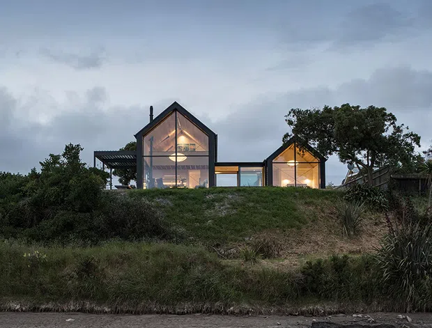 Exterior view of a contemporary residential building at dusk, set behind a grassy and planted embankment. The home is composed of two steeply pitched gabled volumes linked by a lower glazed connector, with extensive floor-to-roof glazing glowing warmly from within. Dark framing, a projecting pergola-like structure, and surrounding coastal-looking vegetation are visible beneath an overcast sky.