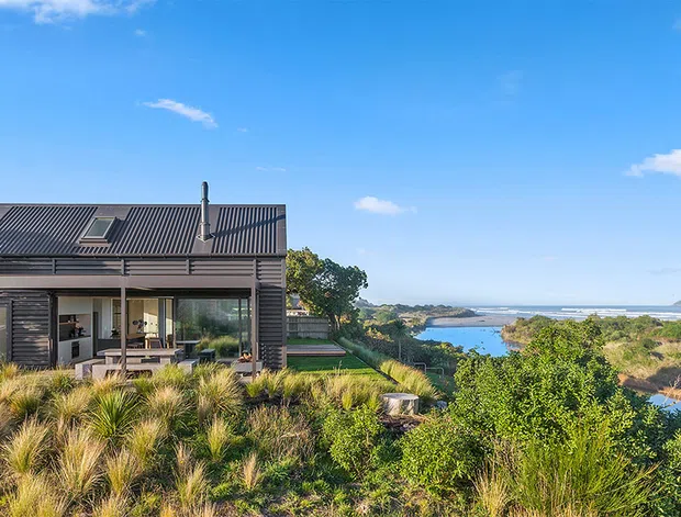 Exterior view of a modern coastal home set within dense native-style landscaping, overlooking a sheltered body of water and distant coastline. The house has dark horizontal cladding, a steep corrugated metal roof with skylights and chimney, extensive glazing, and a covered outdoor living terrace. Bright blue sky and natural grasses reinforce the coastal setting.