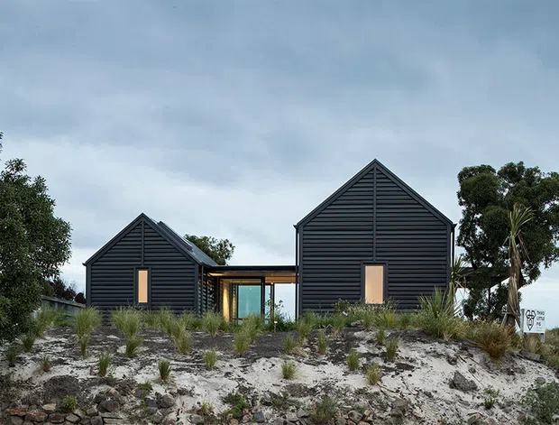 Exterior view of a contemporary coastal-style residence composed of two dark horizontal-clad gabled volumes connected by a glazed central link. The house sits on a sandy, sparsely landscaped site with native grasses and coastal vegetation under an overcast sky. Vertical timber-toned entry or window elements punctuate the dark facades.