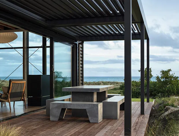 A contemporary coastal outdoor dining terrace beneath a dark slatted pergola, with a substantial concrete dining table and built-in bench seating. Full-height glazed walls reveal a refined interior with timber flooring and a sculptural pendant, while the deck overlooks low coastal vegetation and the ocean. Strong indoor-outdoor connection, dark metal framing, and a relaxed yet architectural atmosphere.