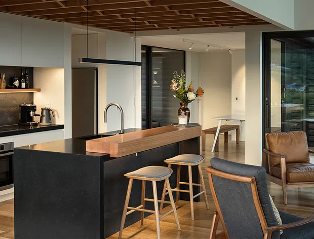A contemporary open-plan kitchen and living area with a large dark island, timber breakfast bar, integrated appliances, and black-framed glazing. Warm timber ceiling battens and flooring contrast with pale cabinetry and dark stone-like surfaces. Timber stools, upholstered lounge chairs, and a leather armchair furnish the adjacent dining and living zone.