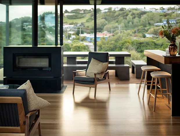 A refined open-plan living and kitchen area with expansive floor-to-ceiling glazing overlooking a leafy suburban landscape. The room features warm timber flooring, a dark built-in fireplace, mid-century-inspired lounge chairs, a streamlined kitchen island with timber cabinetry and a light benchtop, and bar stools. The space is bright, warm, and connected to the surrounding greenery.