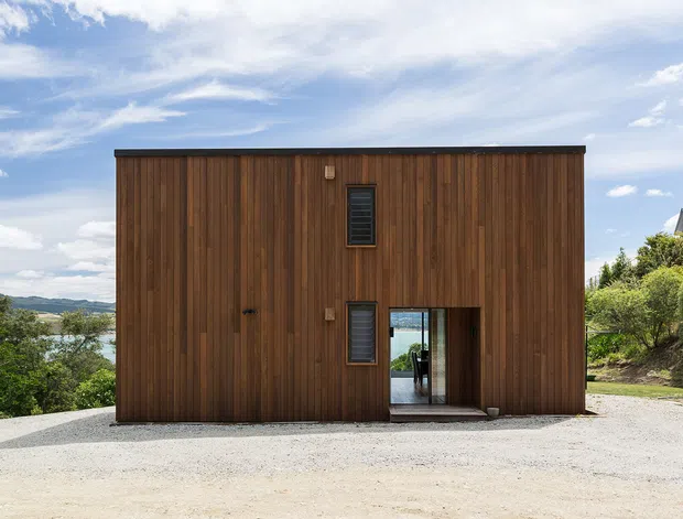 A compact two-storey rectangular building viewed from the exterior, clad almost entirely in vertical dark timber boards. The minimalist facade has narrow dark-framed windows, a small upper-level window, and a recessed glazed entry at ground level. It sits on a pale gravel forecourt with landscaped vegetation and a distant hilly landscape under a partly cloudy sky.