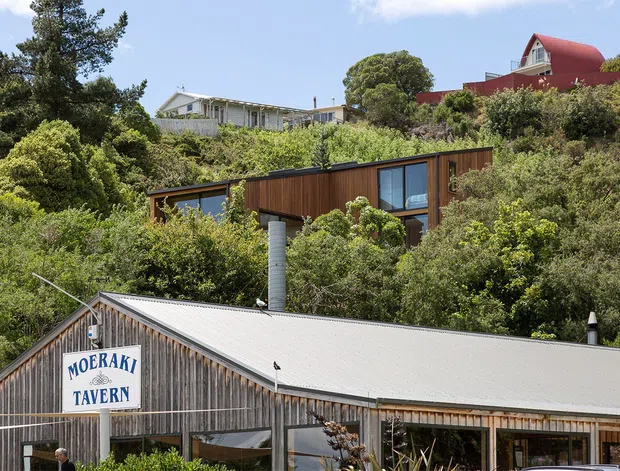 A sunny exterior streetscape showing the Moeraki Tavern in the foreground, with corrugated metal roofing and weathered vertical timber cladding. A modern dark timber-clad house is nestled into dense hillside vegetation behind it, with additional homes visible higher on the slope.