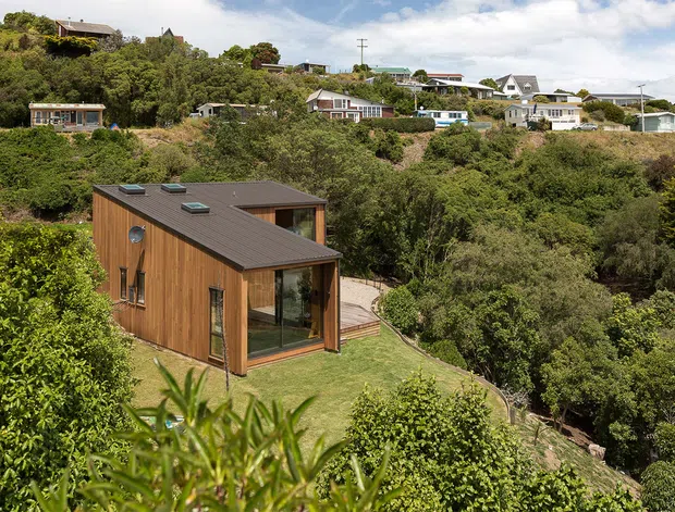 A compact modern timber-clad dwelling set into a densely vegetated, sloping site. The single-storey building has a dark low-pitched roof with several roof skylights, large glazed openings, and a small timber deck overlooking the surrounding bush. Other homes are visible on the hillside in the background.
