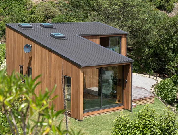 A compact modern timber-clad house set within a leafy landscaped site. The building has a dark low-pitched metal roof with several skylights, warm vertical wood cladding, large glazed openings, and a projecting glazed corner or entry volume. A timber deck and paved path connect the house to the surrounding garden.