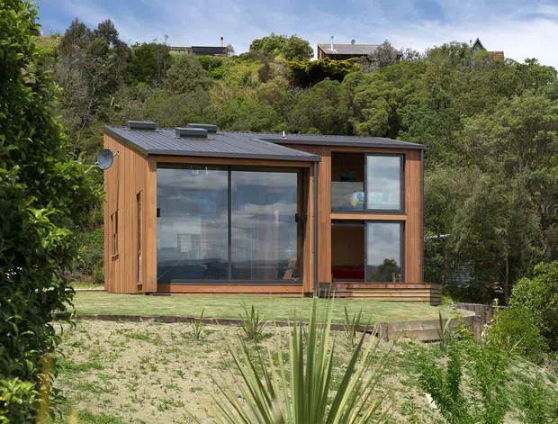 A compact modern timber-clad dwelling set within a lush, wooded landscape. The single-storey structure has a shallow dark metal roof, extensive floor-to-ceiling glazing, a glazed entry or living area, and a projecting two-level glazed section. Low timber decking and sparse coastal planting surround the building.