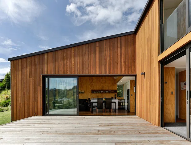 Exterior view of a contemporary timber-clad home opening onto a broad timber deck. The low-pitched asymmetrical roofline, extensive dark-framed glazing, and large open sliding doors connect the deck to a visible dining and kitchen area inside. The surrounding setting includes landscaped lawn and planting beneath a bright sky.