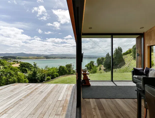A bright contemporary living room opens directly onto a timber deck through expansive floor-to-ceiling glazing, framing panoramic views over a green landscaped site and calm coastal water. The interior features warm timber wall panelling and flooring, a dark-framed glazed wall, a charcoal rug, and dark upholstered seating. The deck and living space create a strong indoor-outdoor connection.