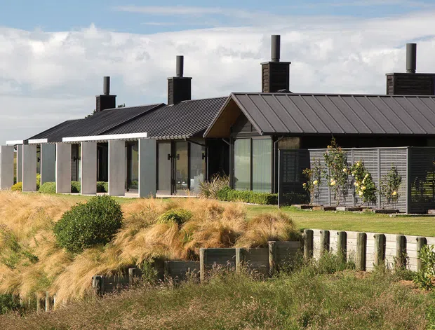 A low, elongated contemporary residential building set in an open rural or coastal landscape. The dark standing-seam pitched roof and dark cladding contrast with pale vertical wall panels and extensive glazing. Multiple chimney flues rise from the roof, while grasses, shrubs, and a low timber retaining structure occupy the foreground.