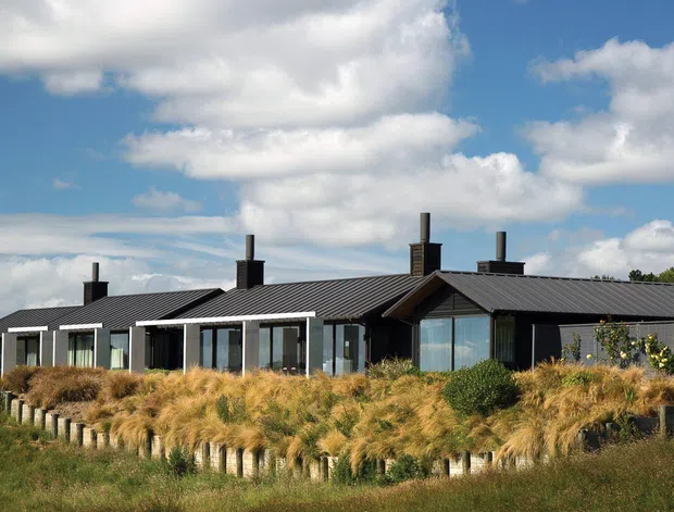 Single-storey contemporary rural residence with a long, low-profile form, dark corrugated metal roofing, extensive floor-to-ceiling glazing, and dark cladding. The house is set within landscaped grasses and a timber-post boundary, under a bright partly cloudy sky.