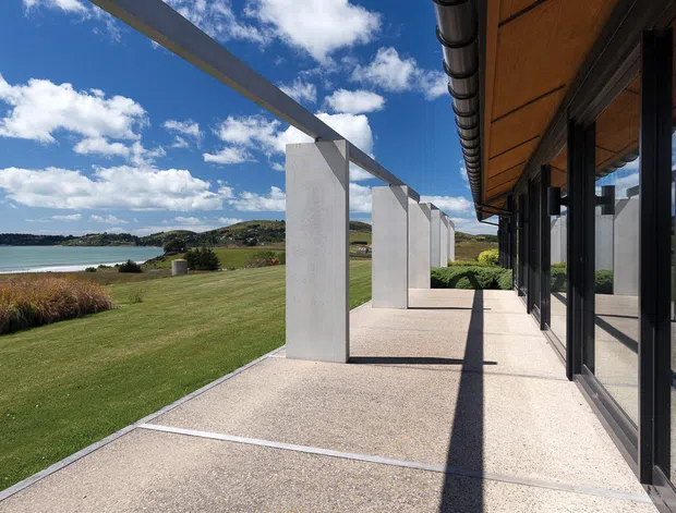 A modern waterfront building viewed from a broad covered terrace or walkway. Full-height glazing with dark frames reflects the landscaped grounds and water, while a warm timber soffit and repeated white structural columns create a strong linear composition. The property sits beside a calm bay with expansive lawn and planted grasses under a bright blue sky.