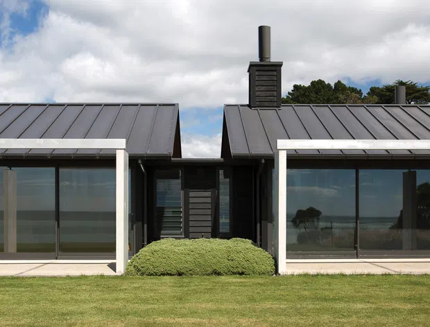 Front exterior view of a low-profile contemporary home composed of dark-clad pavilion-like volumes with simple pitched metal roofs. Large glazed openings are framed in white, with covered recessed verandah areas, chimney elements, a central entry connection, and a broad lawn in the foreground. The image shows a rural or open landscape beyond the glazing, under a partly cloudy sky.
