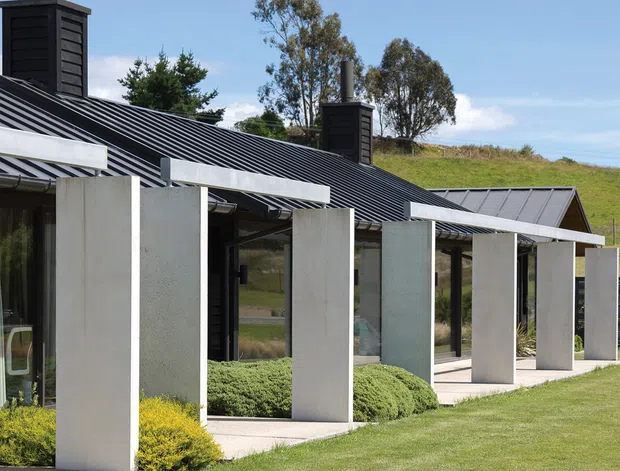 Contemporary single-storey building exterior with dark horizontal cladding, long pitched metal roofs, tall black chimneys, and a covered verandah defined by repeated pale concrete or rendered piers. Floor-to-ceiling glazing sits behind the colonnade, opening toward a neatly landscaped lawn and rolling rural grassland.