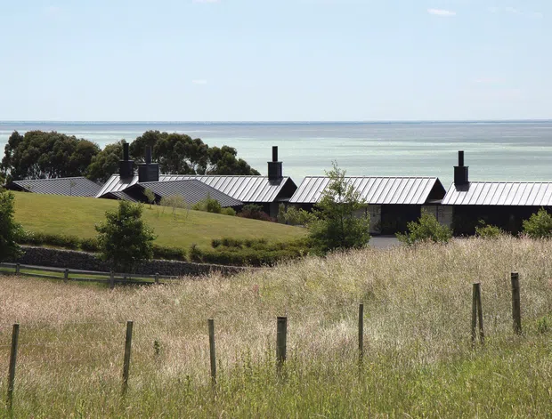 A low, elongated residential building with several connected sections and repeated pitched metal roofs sits in a grassy coastal landscape overlooking a broad body of water. Dark chimneys punctuate the roofline, while mature trees and rolling meadow surround the structure. The image has a quiet rural-coastal character with simple contemporary forms.